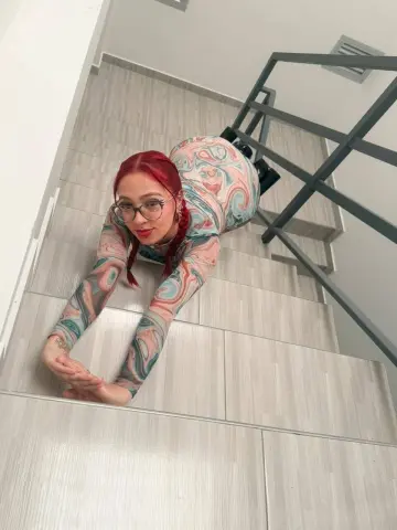 amelie_adams poses on a tiled floor wearing a long-sleeved, swirl-patterned bodysuit and glasses, looking up at the camera with red hair.