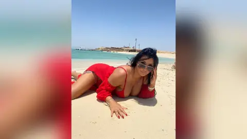 MissyJolie lies on the sand wearing a red dress and sunglasses, looking toward the camera with her hair down.