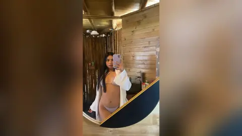 Young woman in a white robe and bikini taking a mirror selfie in a rustic wooden tropical villa.