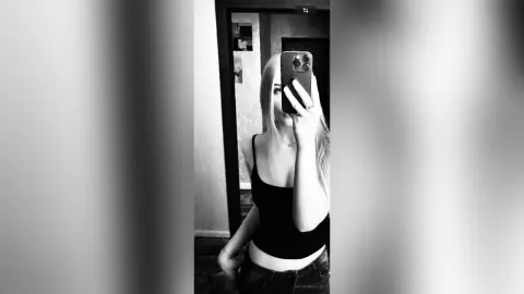 KathlineVelzy poses for a mirror selfie wearing a black tank top and dark jeans, showing her long blonde hair and slim build.