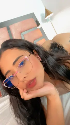 Young woman with long dark hair and pink glasses relaxing in a cozy bedroom setting.