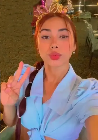 Young woman in a light blue blouse wearing a gold headband making a peace sign at an outdoor evening party.
