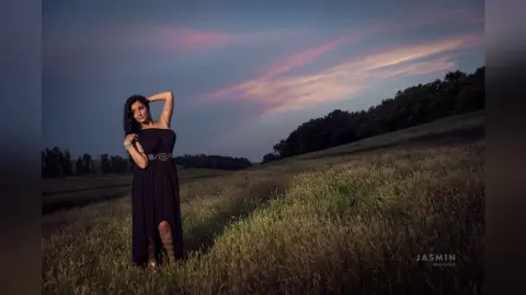 MissyJolie stands in a grassy field at dusk wearing a black strapless dress with a gold decorative belt.