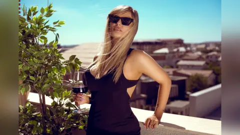 Gia poses on a balcony wearing a black tank top and sunglasses, holding a glass of red wine.