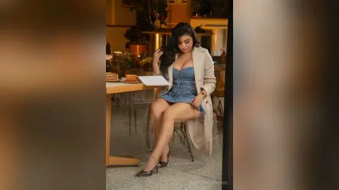 Elegant woman in a blue dress and beige coat sitting at a cafe table with a relaxed, sophisticated mood.