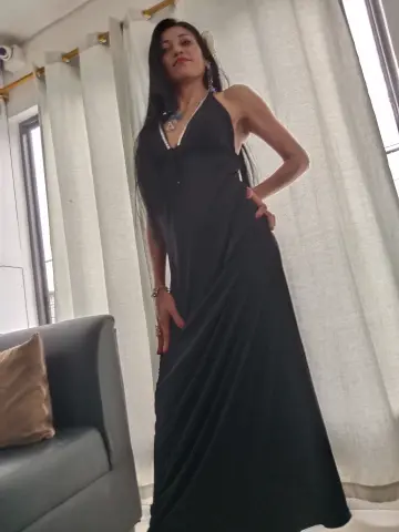 Elegant woman in a long black halter dress standing by a window in a bright, modern living room.