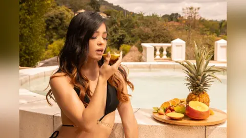 Young woman in a black bikini eating fruit by a luxury poolside on a sunny summer day.