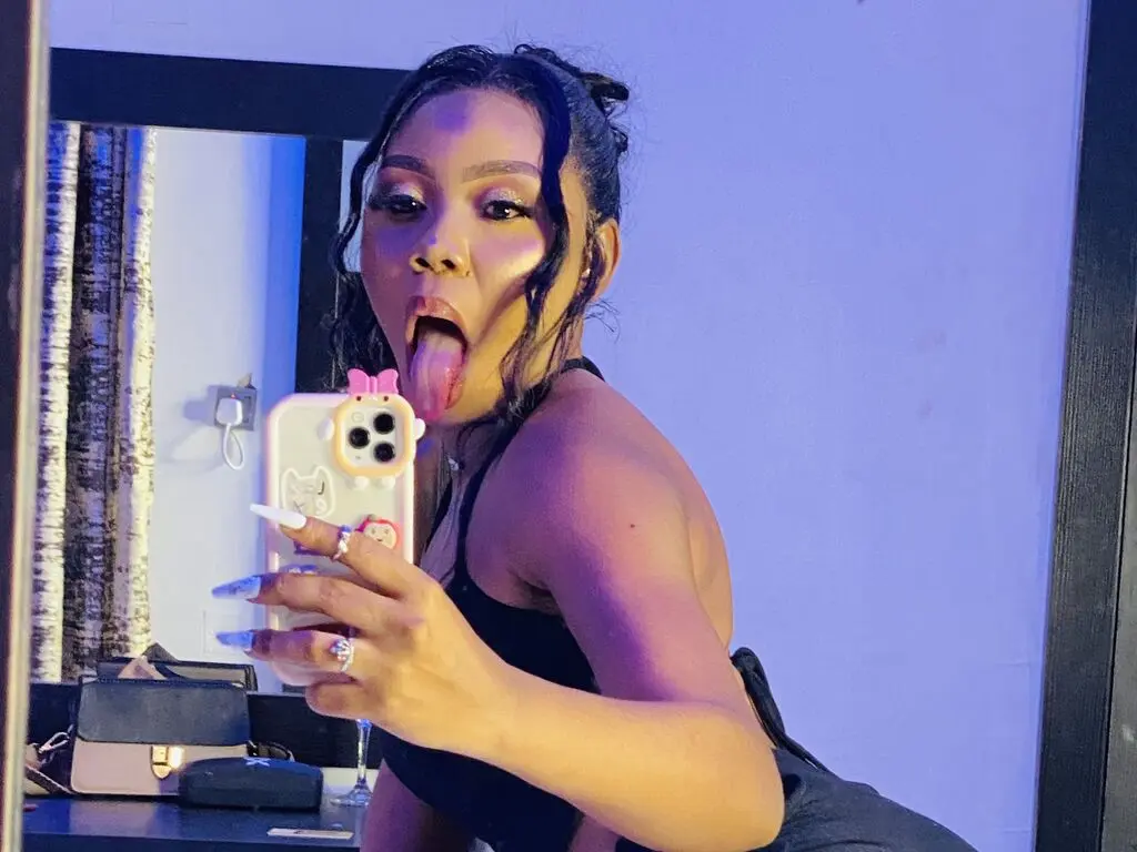 Young woman in black tank top taking a playful selfie in a mirror with warm indoor lighting.
