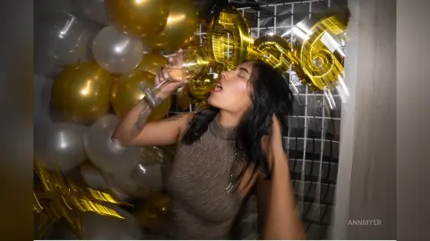 Young woman in a sparkly top celebrating a birthday with gold balloons and champagne in a festive mood.