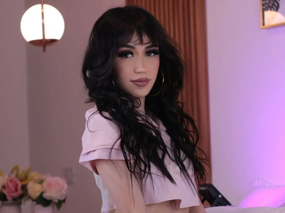 ThaliaZafirr poses with long wavy black hair, wearing a pink cleavage-baring top against a plain wall background.