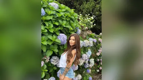 ReynaGomes poses outdoors with long wavy hair, wearing a white tank top and denim shorts against a backdrop of green leaves and purple hydrangeas.