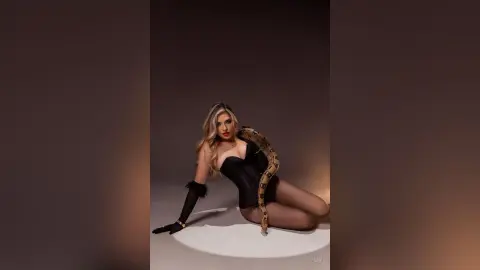 Glamorous woman in a black bodysuit and long gloves posing with a large snake in a moody studio setting.