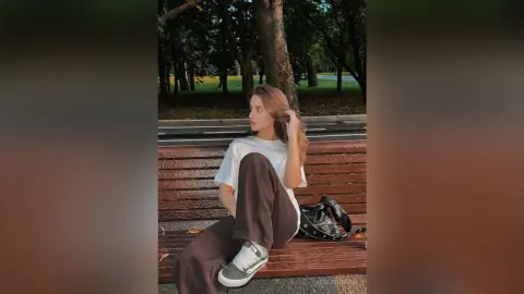 LeslieDorian sits on a park bench wearing a white t-shirt and dark pants, looking away with her long hair flowing.