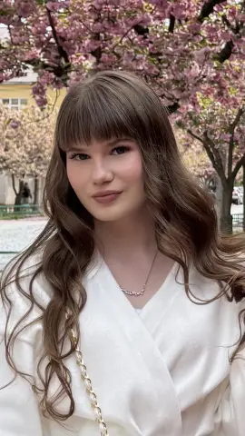 AshleyFierce poses outdoors with long wavy brown hair and bangs, wearing a white wrap top and a delicate silver necklace against a background of pink cherry blossoms.