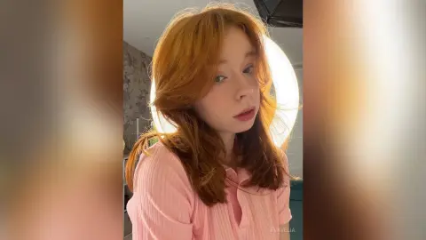 Redheaded woman in a pink ribbed shirt posing under warm ring light in a cozy indoor setting.