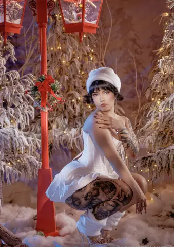 Tattooed woman in a white dress posing in a snowy winter wonderland setting with festive red lampposts.
