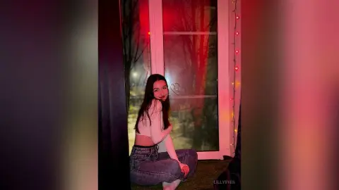 Young woman in a white long-sleeve crop top and jeans sitting by a window with moody red lighting.