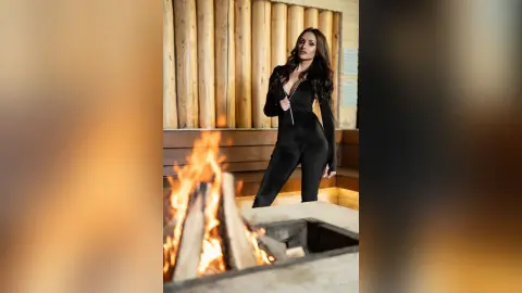 Elegant woman in a black long-sleeve jumpsuit posing by a glowing outdoor fire pit in a rustic log cabin setting.