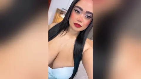 NiaCorelli poses in a white top, showing her cleavage and long black hair with heavy eye makeup and red lipstick.