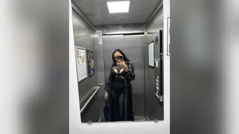 NicoleBella stands in an elevator wearing a black cut-out bodysuit and a long black robe, holding a phone for a mirror selfie.