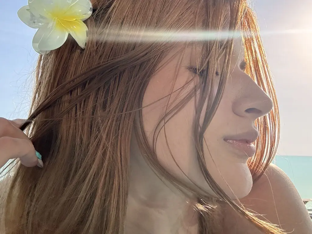 Close-up of a woman with long brown hair and a white flower in a sunny, tropical outdoor setting.