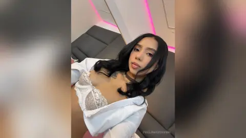 Young woman with long dark hair in a white lace top relaxing on a grey sofa in a room with pink neon lighting.