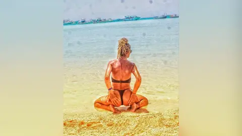 DelliaSmith sits on a sandy beach facing the ocean, wearing a black bikini and showing her bare back and buttocks while touching her bottom.