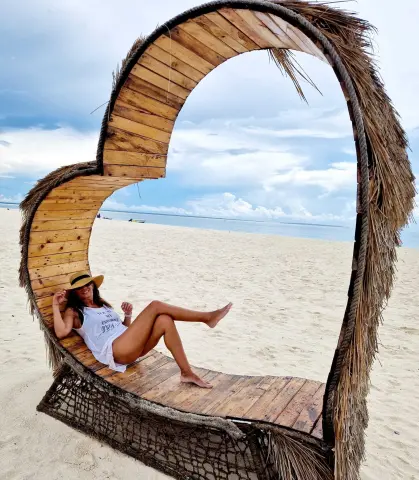 Relaxed woman in a white tank top and sun hat lounging in a heart-shaped wooden beach chair under a cloudy sky.