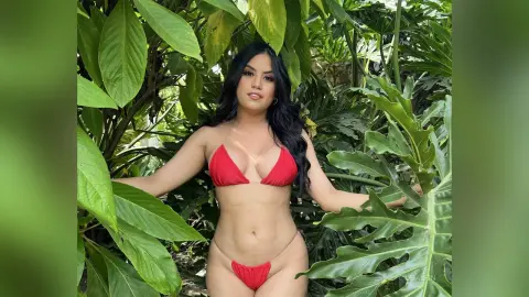 LeiaLein — Young woman in a red bikini posing amidst lush tropical green leaves in a sunny garden setting.