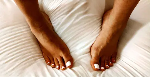Close-up of bare feet resting on white wrinkled bed sheets in a bright, cozy bedroom setting.