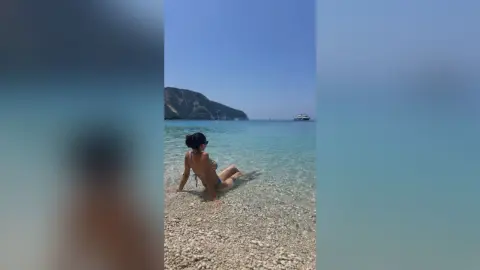 Woman in a bikini relaxing on a sunny pebble beach by turquoise ocean water with mountains in the background.