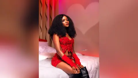 Glamorous woman in a red sequin crop top and matching shorts sitting on a bed against a romantic heart backdrop.