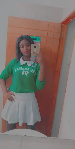 Young woman in a green Los Angeles sweatshirt and white pleated skirt taking a mirror selfie in a bathroom.