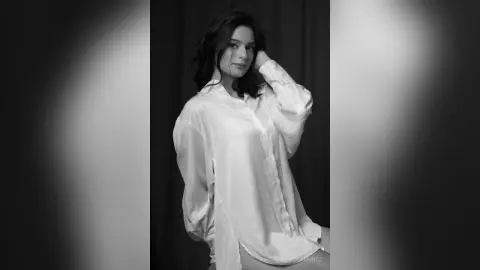 Moody black and white portrait of a woman in a white oversized button-down shirt against a dark background.