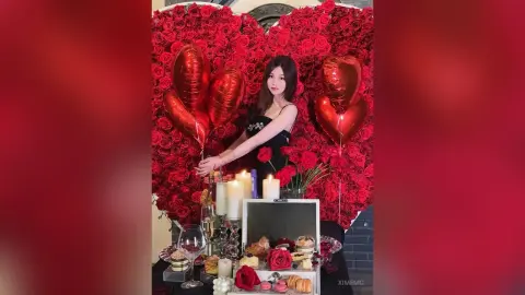Elegant woman in a black dress posing by a romantic red rose wall and heart balloons for a Valentine's celebration.