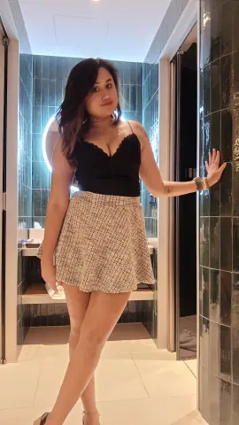 Stylish woman in a black lace camisole and tweed mini skirt posing in a modern blue tiled bathroom.