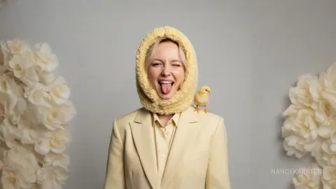 NancyKarsten — Blonde woman in a cream blazer and fuzzy hood playfully sticking her tongue out next to a small yellow chick.