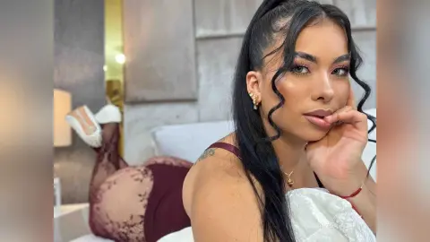 VictoriaCapri poses with long black hair and dark eye makeup, wearing a white lace top and maroon lace leggings while lying on a bed.