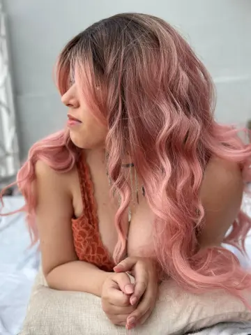 Izzumi poses with her long, wavy pink hair falling over her shoulder while wearing an orange lace bra.