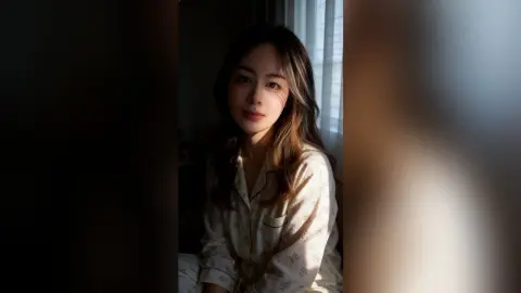 Young woman in cream patterned pajamas relaxing in soft, warm sunlight by a window.
