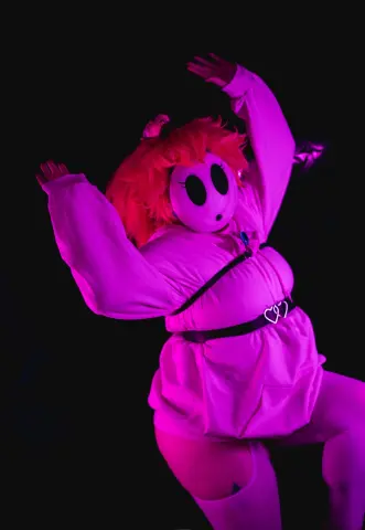 MagicalMae wears a pink costume and a white mask while posing against a dark background under pink lighting.