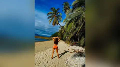 Woman in orange bikini walking on a tropical sandy beach lined with palm trees under a blue sky.