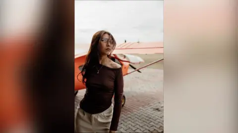 Stylish woman in a dark long-sleeve top and sunglasses posing by a vintage orange airplane on a cloudy day.