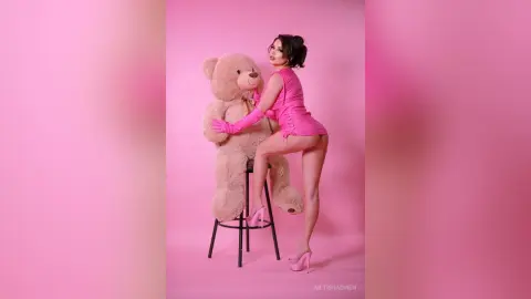Glamorous woman in a pink mini dress and heels posing with a giant teddy bear against a pink studio background.