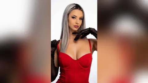 Kalira poses in a red corset and black gloves, showing off her long silver hair and cleavage against a white background.