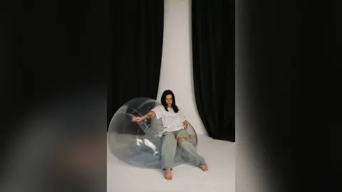 Young woman in casual white shirt and grey trousers sitting inside a large bubble in a minimalist studio.