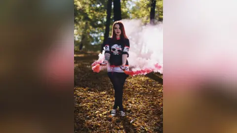 Red-haired woman in a black graphic t-shirt holding pink smoke bombs in a moody autumn forest setting.