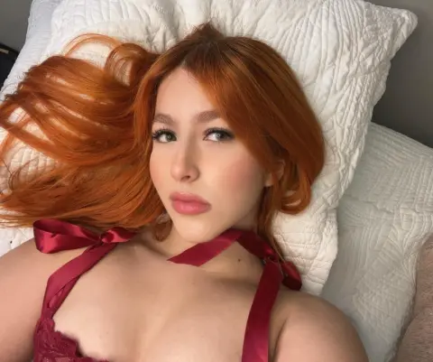 Redheaded woman in a burgundy lace lingerie set relaxing on white pillows with a dreamy mood.