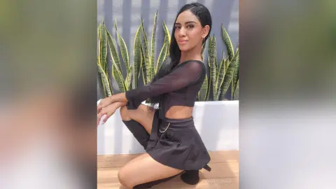 Young woman in a black long-sleeve crop top and mini skirt posing confidently in front of indoor plants.