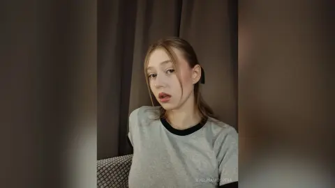 Young woman in a grey crewneck sweater posing with a moody expression against dark curtains.